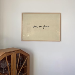 May include: A framed print with the words "vino, per favore" in black script on a cream-colored background. The frame is a light wood color. The print is hanging on a white wall.