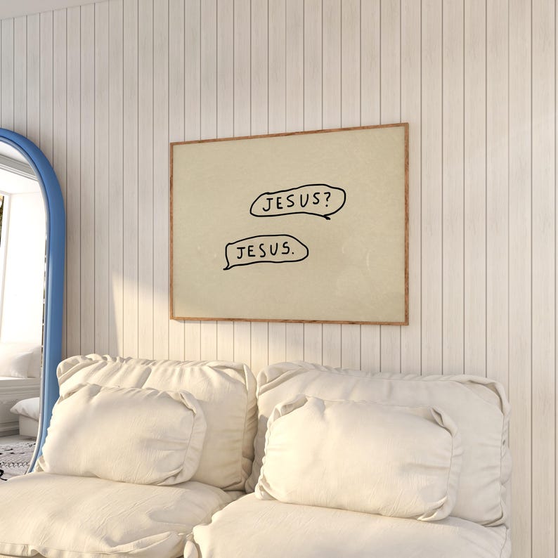 May include: A framed print with the text "JESUS?" and "JESUS." in black speech bubbles. The artwork has a beige background and a brown frame. The print hangs on a white paneled wall above a white sofa with pillows.