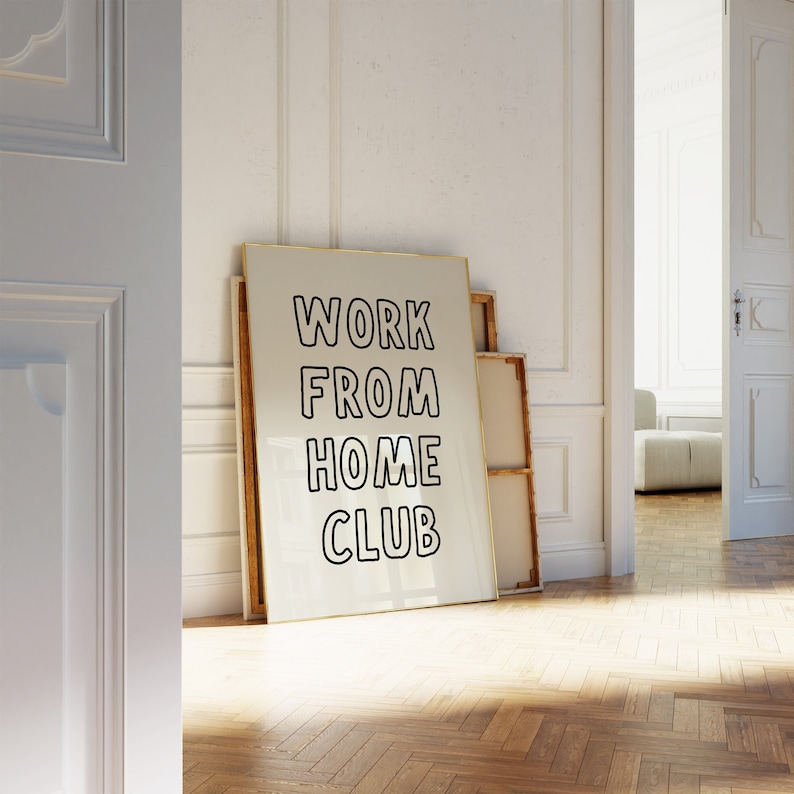 Op de afbeelding: Een ingelijste poster met de woorden "WORK FROM HOME CLUB" in vette zwarte letters. De poster leunt tegen een witte muur met andere ingelijste kunstwerken. De kamer heeft een parketvloer en een open deur.