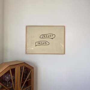 May include: Framed artwork with the words "JESUS?" and "JESUS." in black speech bubbles on a beige background. The artwork is in a light brown wooden frame, hanging on a white wall.