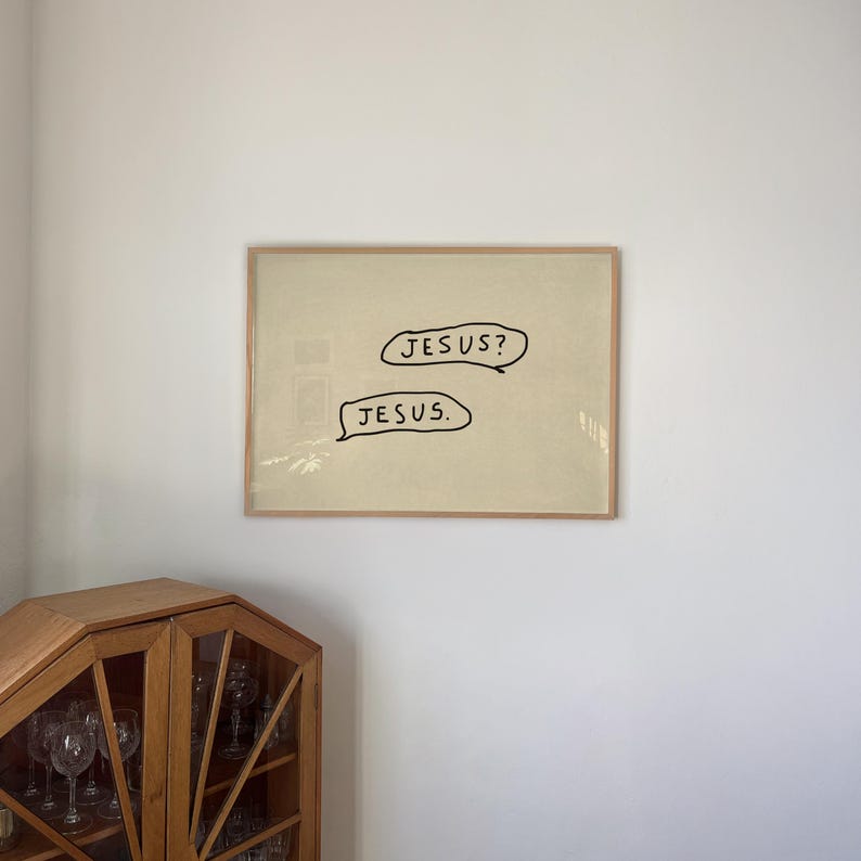 May include: Framed artwork with the words "JESUS?" and "JESUS." in black speech bubbles on a beige background. The artwork is in a light brown wooden frame, hanging on a white wall.