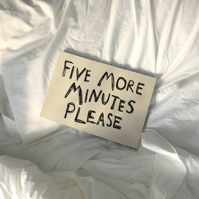 May include: A rectangular piece of tan paper with the handwritten text "FIVE MORE MINUTES PLEASE" in black ink. The paper rests on a bed of white sheets and pillows, creating a soft, inviting background.