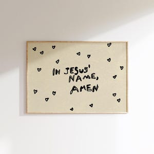 May include: A framed print with the words "IN JESUS' NAME, AMEN" in black, handwritten-style font. Small black hearts are scattered around the text on a beige background. The frame is a light brown color.