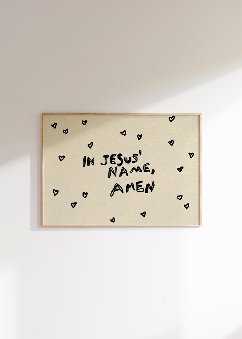 May include: A framed print with the words "IN JESUS' NAME, AMEN" in black, handwritten-style font. Small black hearts are scattered around the text on a beige background. The frame is a light brown color.