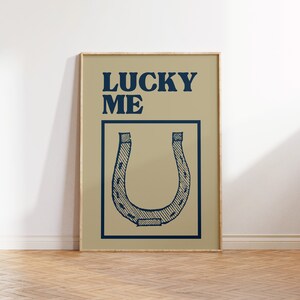 Puede incluir: Un p&oacute;ster beige con marco de madera que presenta una herradura azul y el texto "LUCKY ME" en azul.
