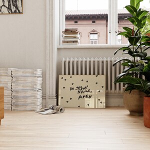 May include: A framed artwork with the text "IN JESUS' NAME, AMEN" in black lettering, surrounded by small heart shapes. The artwork is displayed on a light-colored wooden floor, near a window and potted plants.