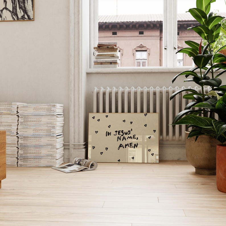 May include: A framed artwork with the text "IN JESUS' NAME, AMEN" in black lettering, surrounded by small heart shapes. The artwork is displayed on a light-colored wooden floor, near a window and potted plants.