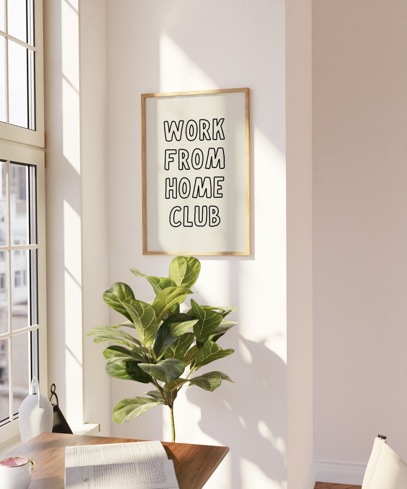 Op de afbeelding: Een ingelijste print met de tekst "WORK FROM HOME CLUB" in zwarte letters. De print zit in een licht houten frame en hangt aan een witte muur. Een groene plant staat eronder, bij een raam.
