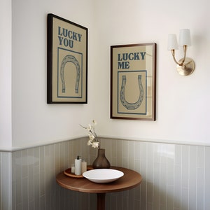 Puede incluir: Dos impresiones enmarcadas con un dise&ntilde;o de herradura y el texto "Lucky You" y "Lucky Me" colgadas en una pared blanca. Una l&aacute;mpara de pared blanca y dorada con dos bombillas est&aacute; en el lado derecho de la imagen. Una mesa de madera con un taz&oacute;n blanco y un jarr&oacute;n con flores blancas est&aacute; en la esquina inferior izquierda de la imagen.