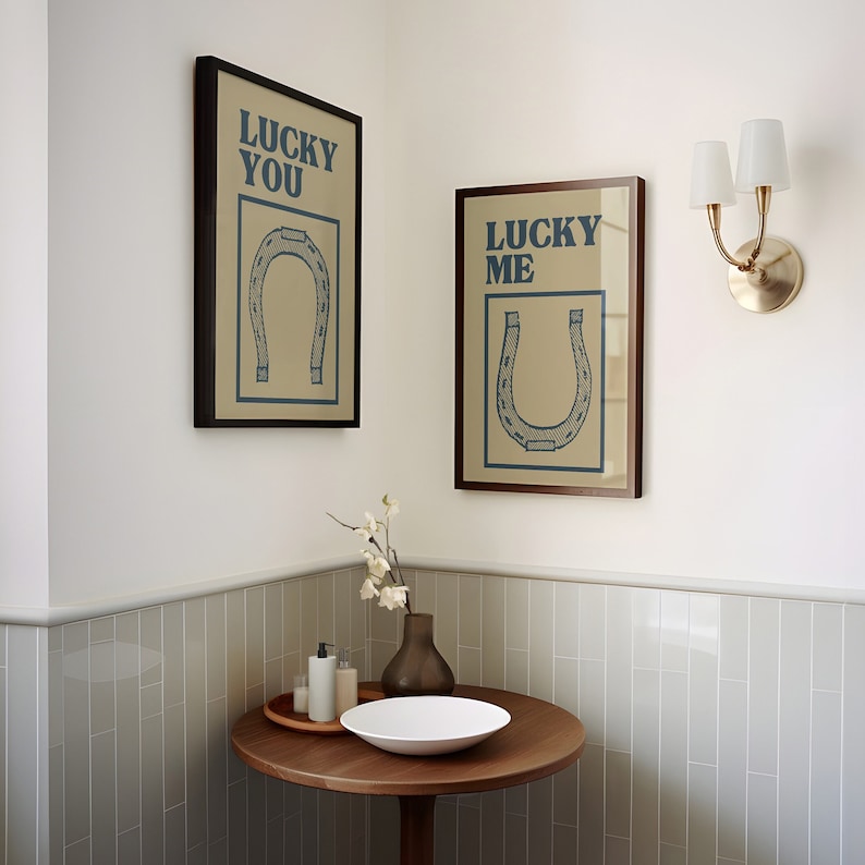 Puede incluir: Dos impresiones enmarcadas con un dise&ntilde;o de herradura y el texto "Lucky You" y "Lucky Me" colgadas en una pared blanca. Una l&aacute;mpara de pared blanca y dorada con dos bombillas est&aacute; en el lado derecho de la imagen. Una mesa de madera con un taz&oacute;n blanco y un jarr&oacute;n con flores blancas est&aacute; en la esquina inferior izquierda de la imagen.