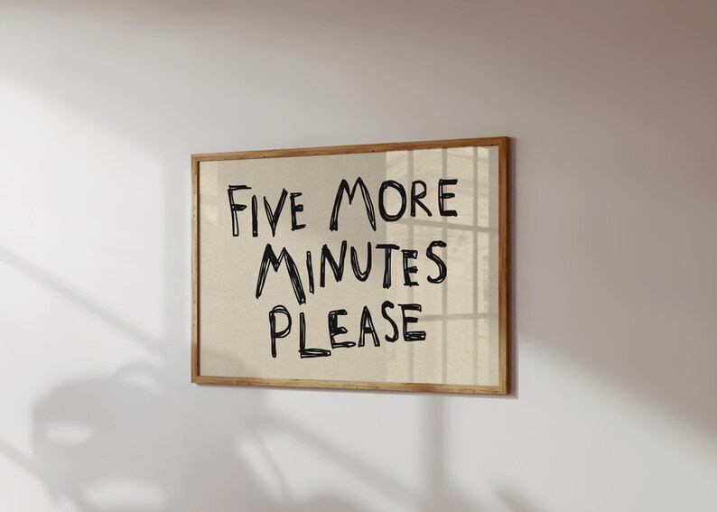 May include: A framed wall art print with the text "FIVE MORE MINUTES PLEASE" in black handwritten style on a beige background. The frame is made of light brown wood. The artwork is mounted on a white wall.