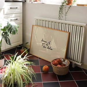 May include: A framed print with the text "kisses if you do the dishes" leans against a radiator. A basket of apples and a single peach sit on a red and black tiled floor. Plants and a cookbook are also visible.
