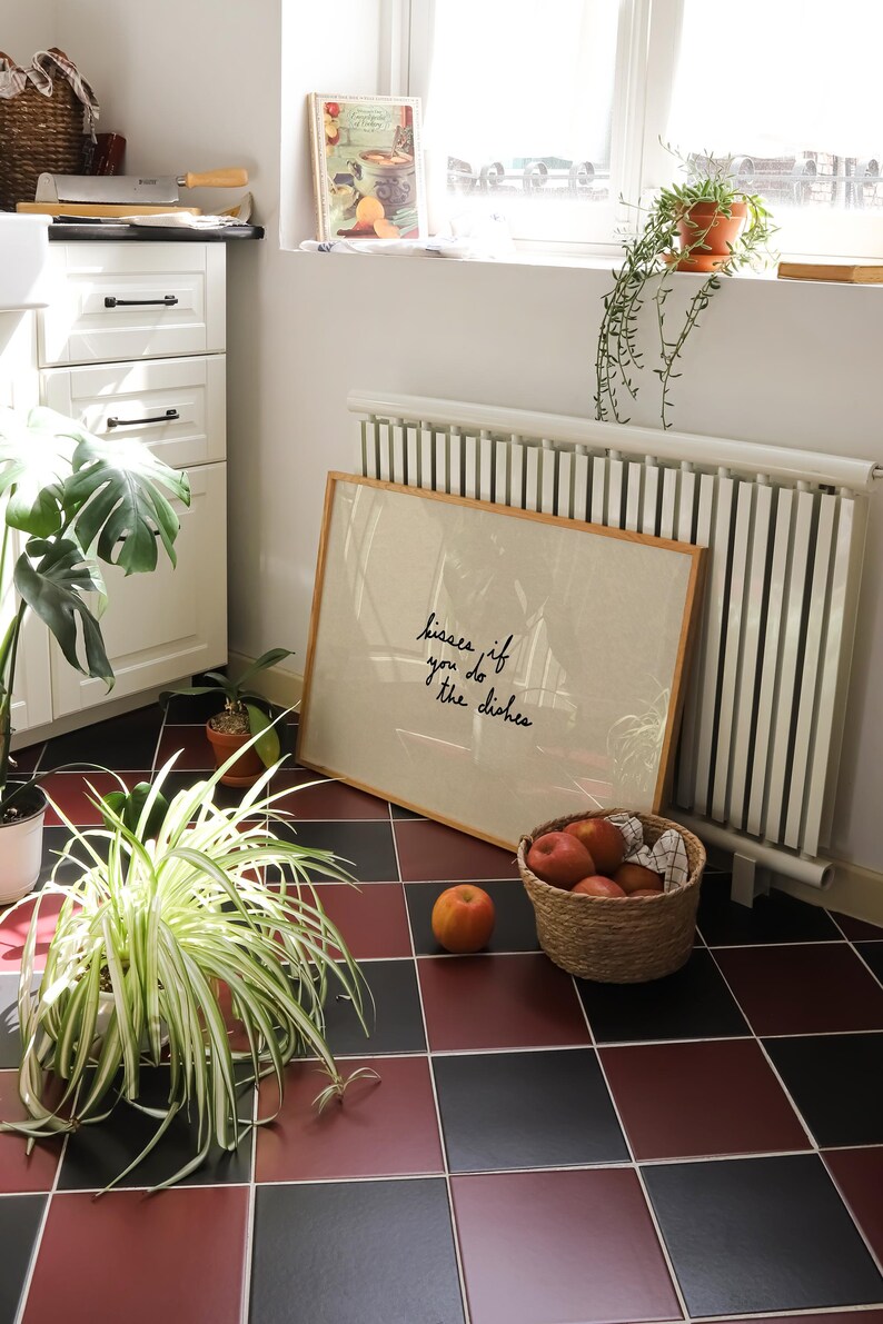 May include: A framed print with the text "kisses if you do the dishes" leans against a radiator. A basket of apples and a single peach sit on a red and black tiled floor. Plants and a cookbook are also visible.