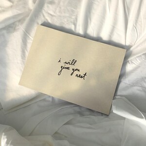 Puede incluir: Una tarjeta beige con el texto manuscrito "i will give you rest" en escritura negra. La tarjeta descansa sobre una cama de s&aacute;banas blancas arrugadas, creando una est&eacute;tica suave y relajante.