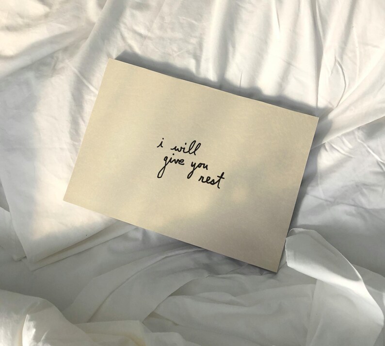 Puede incluir: Una tarjeta beige con el texto manuscrito "i will give you rest" en escritura negra. La tarjeta descansa sobre una cama de s&aacute;banas blancas arrugadas, creando una est&eacute;tica suave y relajante.