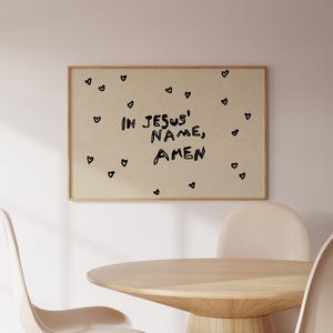 May include: A framed print with the words "IN JESUS' NAME, AMEN" in black lettering, surrounded by small black heart shapes. The print hangs on a light-colored wall above a wooden table and chairs.