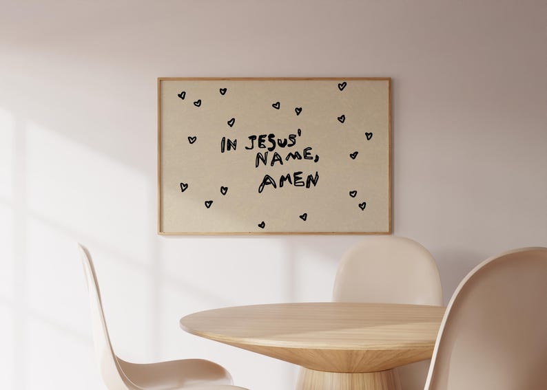 May include: A framed print with the words "IN JESUS' NAME, AMEN" in black lettering, surrounded by small black heart shapes. The print hangs on a light-colored wall above a wooden table and chairs.