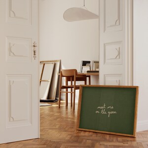 May include: A framed chalkboard with the text "meet me on the green" in white script. The chalkboard has a dark green surface and a light brown wooden frame. The image is taken indoors, with a wooden floor and a partially visible room in the background.