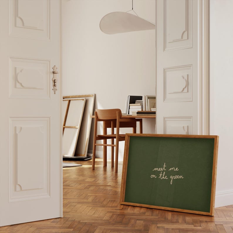 May include: A framed chalkboard with the text "meet me on the green" in white script. The chalkboard has a dark green surface and a light brown wooden frame. The image is taken indoors, with a wooden floor and a partially visible room in the background.