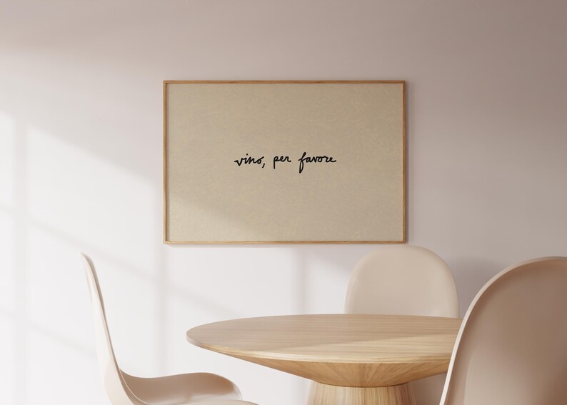 May include: A framed print with the words "vino, per favore" in black script. The print is displayed above a wooden table and chairs, creating a dining room scene. The frame is a light brown color.