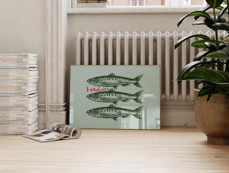 May include: A framed print featuring three green fish illustrations with the word "Sardines" in red. The artwork is displayed on a light green background, leaning against a wall. The print is in a room with a plant and a stack of magazines.
