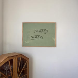 May include: A framed artwork with a light green background and the words "MARGS?" and "MARGS." in black speech bubbles. The frame is a light brown color. The artwork is hanging on a white wall.