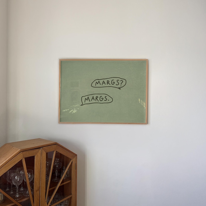 May include: A framed artwork with a light green background and the words "MARGS?" and "MARGS." in black speech bubbles. The frame is a light brown color. The artwork is hanging on a white wall.