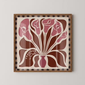 May include: A square framed wall art print featuring a stylized illustration of pink calla lilies in a pink vase against a cream background. The artwork includes the text "CONSIDER THE LILIES" and is bordered by a dark brown and cream checkered pattern.