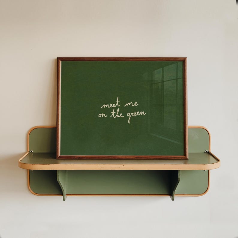 May include: A framed green chalkboard with the text "meet me on the green" in white script. The frame is brown. The chalkboard rests on a two-tiered green and beige shelf, mounted on a white wall.