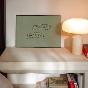 May include: A framed artwork with the text "MARGS?" and "MARGS." in speech bubbles. A white mushroom-shaped lamp and a red book are on a white shelf. A wooden bookshelf is visible on the left.