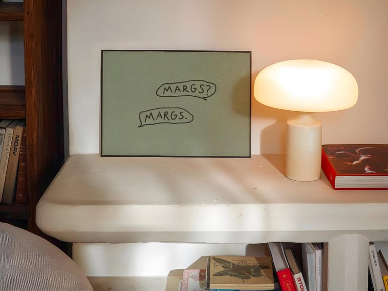 May include: A framed artwork with the text "MARGS?" and "MARGS." in speech bubbles. A white mushroom-shaped lamp and a red book are on a white shelf. A wooden bookshelf is visible on the left.