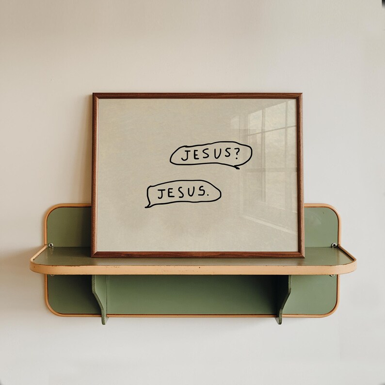 May include: A framed print with the text "JESUS?" and "JESUS." in speech bubbles. The artwork is displayed on a green and wood-toned shelf, mounted on a white wall. The frame is a dark brown color.