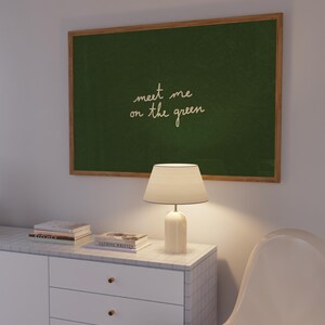 May include: A framed green chalkboard with the text "meet me on the green" hangs above a white cabinet. A white lamp with a beige lampshade sits on the cabinet, next to a stack of books. A white chair is in the foreground.