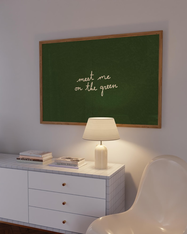May include: A framed green chalkboard with the text "meet me on the green" hangs above a white cabinet. A white lamp with a beige lampshade sits on the cabinet, next to a stack of books. A white chair is in the foreground.