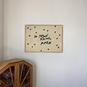 May include: A framed artwork with the text "IN JESUS' NAME, AMEN" in black lettering, surrounded by small black heart illustrations. The artwork is set against a cream-colored background and framed in light wood.