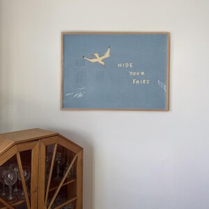 May include: A framed art print featuring a light blue background with a flying seagull illustration. The text "HIDE YOUR FRIES" is written in white. The frame is made of light brown wood. A wooden cabinet is in the lower left corner.