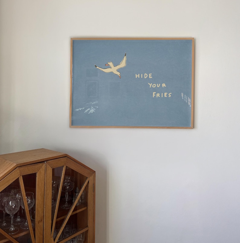 May include: A framed art print featuring a light blue background with a flying seagull illustration. The text "HIDE YOUR FRIES" is written in white. The frame is made of light brown wood. A wooden cabinet is in the lower left corner.