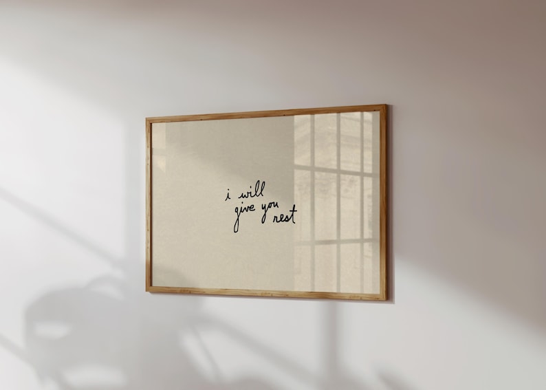 Puede incluir: Un estampado enmarcado con el texto manuscrito "i will give you rest" en escritura negra. La obra de arte tiene un fondo beige y un marco de madera natural. El estampado est&aacute; montado en una pared blanca.