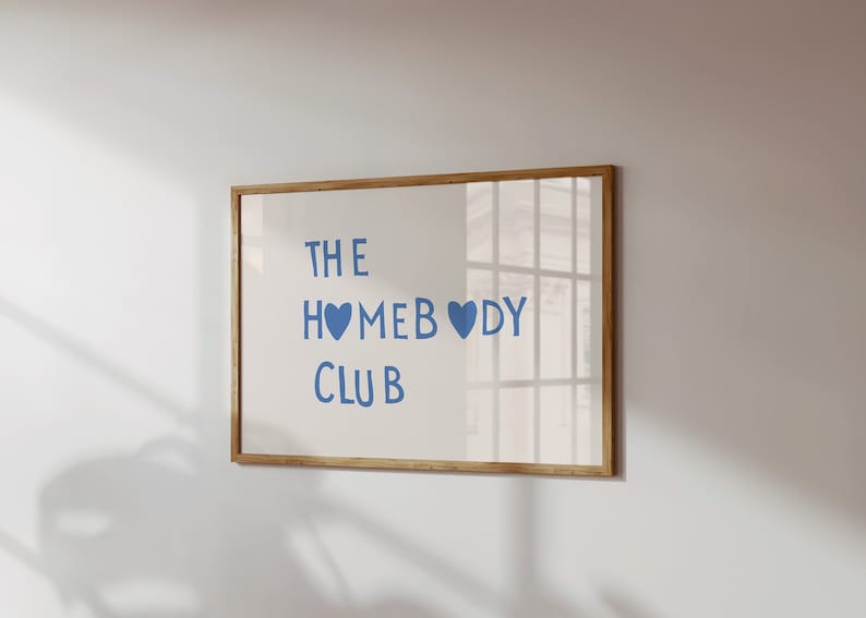 May include: A framed print with the text 'THE HOMEBODY CLUB' in blue lettering with two blue hearts. The print is in a wooden frame and is hanging on a white wall.