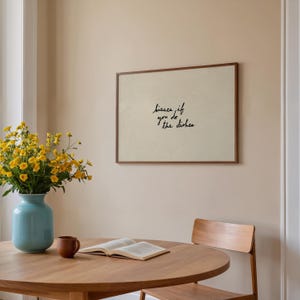 May include: A framed print with the text "kisses if you do the dishes" in black script. The print hangs on a beige wall above a wooden table. A blue vase with yellow flowers and a brown mug are on the table.