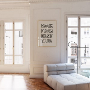 Op de afbeelding: Een ingelijste print met de tekst "WORK FROM HOME CLUB" hangt aan een witte muur. De kamer heeft een lichtgrijze chaise longue, grote ramen en een zwarte vloerlamp. De algehele esthetiek is modern en minimalistisch.