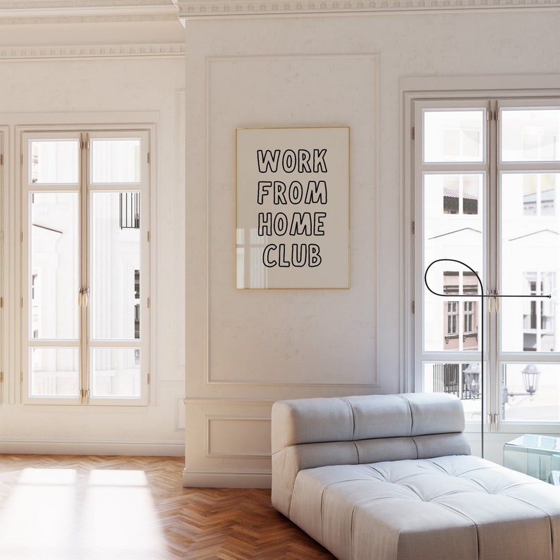 Op de afbeelding: Een ingelijste print met de tekst "WORK FROM HOME CLUB" hangt aan een witte muur. De kamer heeft een lichtgrijze chaise longue, grote ramen en een zwarte vloerlamp. De algehele esthetiek is modern en minimalistisch.