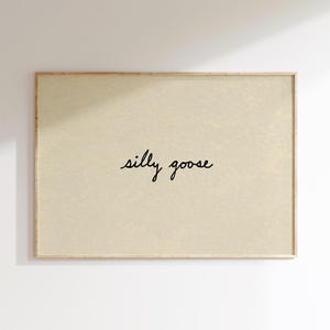 May include: A framed art print with a light beige background. The words "silly goose" are written in a cursive font in the center of the print. The frame is a light brown color.
