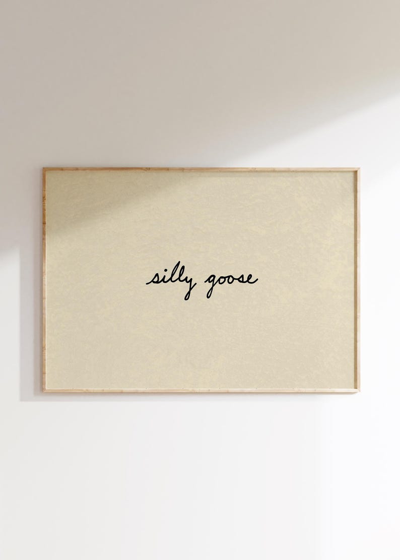 May include: A framed art print with a light beige background. The words "silly goose" are written in a cursive font in the center of the print. The frame is a light brown color.