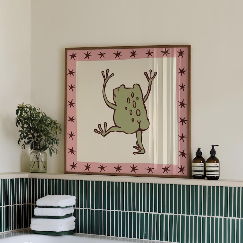 May include: A square framed wall art print featuring a light green cartoon frog with its arms raised and one leg lifted. The illustration is centred on a cream background, surrounded by a pink border decorated with dark brown star shapes.