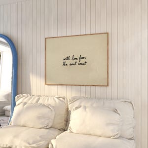 Puede incluir: Un estampado enmarcado con el texto "with love from the east coast" en escritura negra. El estampado tiene un fondo beige y un marco marr&oacute;n. El estampado est&aacute; colgado en una pared blanca sobre un sof&aacute; blanco con cojines blancos.