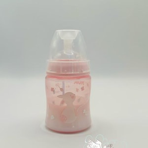 Op de afbeelding: Een roze babyfles met een doorzichtige bovenkant en een roze basis. De fles heeft een cartoon konijn en bloemen ontwerp. Op de fles staat het woord "NUK".