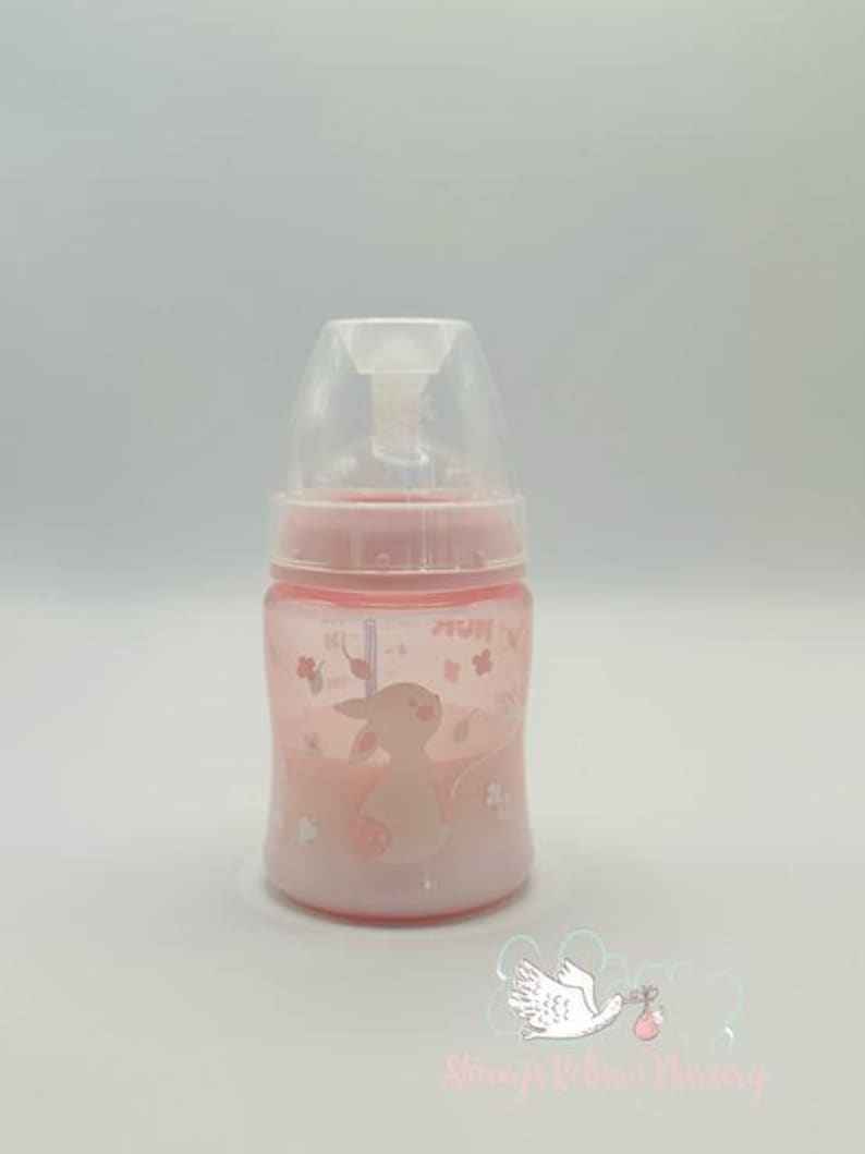 Op de afbeelding: Een roze babyfles met een doorzichtige bovenkant en een roze basis. De fles heeft een cartoon konijn en bloemen ontwerp. Op de fles staat het woord "NUK".