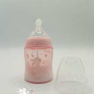 Op de afbeelding: Een roze babyfles met een doorzichtige speen en een bijpassende dop. De fles heeft een cartoon dierenontwerp en bloemaccenten. De fles staat naast een doorzichtige plastic dop.