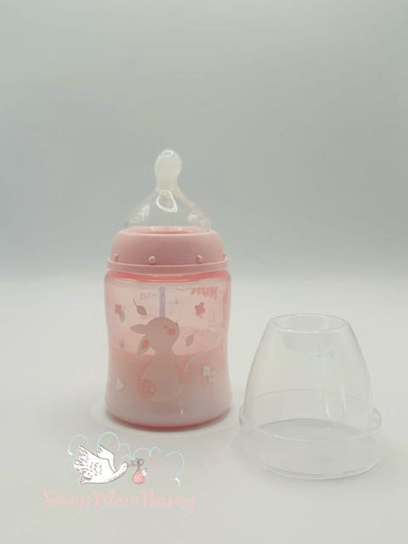 Op de afbeelding: Een roze babyfles met een doorzichtige speen en een bijpassende dop. De fles heeft een cartoon dierenontwerp en bloemaccenten. De fles staat naast een doorzichtige plastic dop.